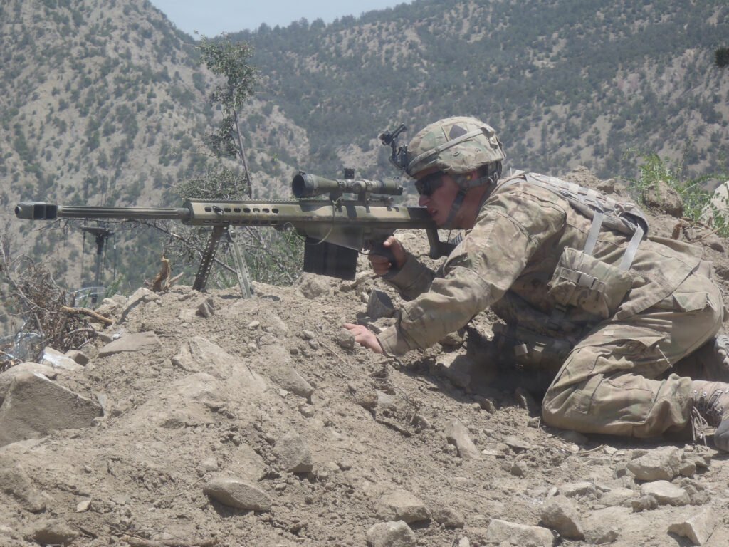 U.S. Army soldier in camouflage lying prone on a rocky ridge, sighting a long-range rifle