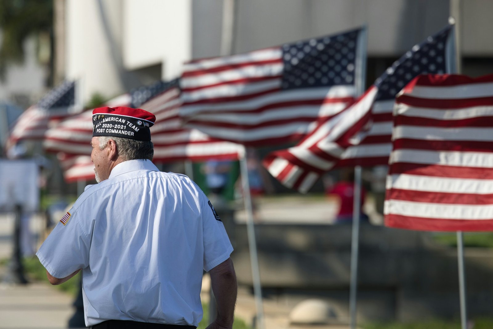 An older veteran carrying a large American flag over his shoulder, walking past a display of multiple flags outdoors.