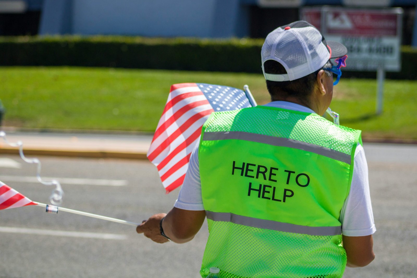 A person wearing a bright yellow vest with 'Here to Help' written on the back, holding an American flag outdoors.