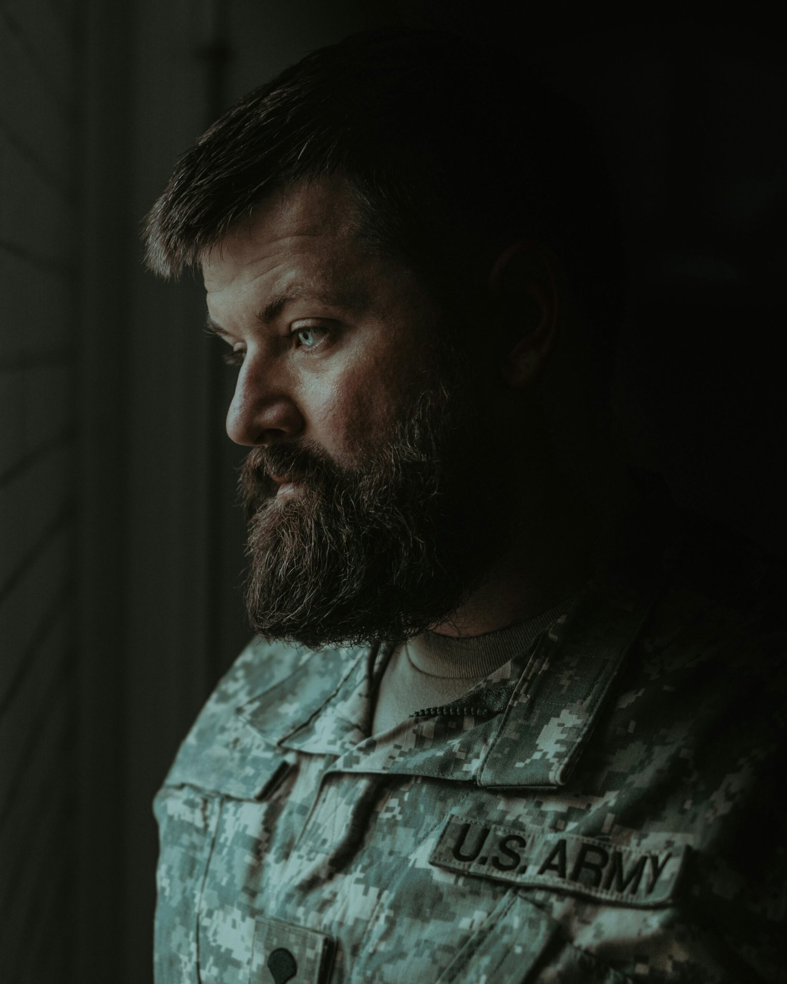 A moody profile portrait of a bearded veteran in a camouflage uniform looking downward thoughtfully.