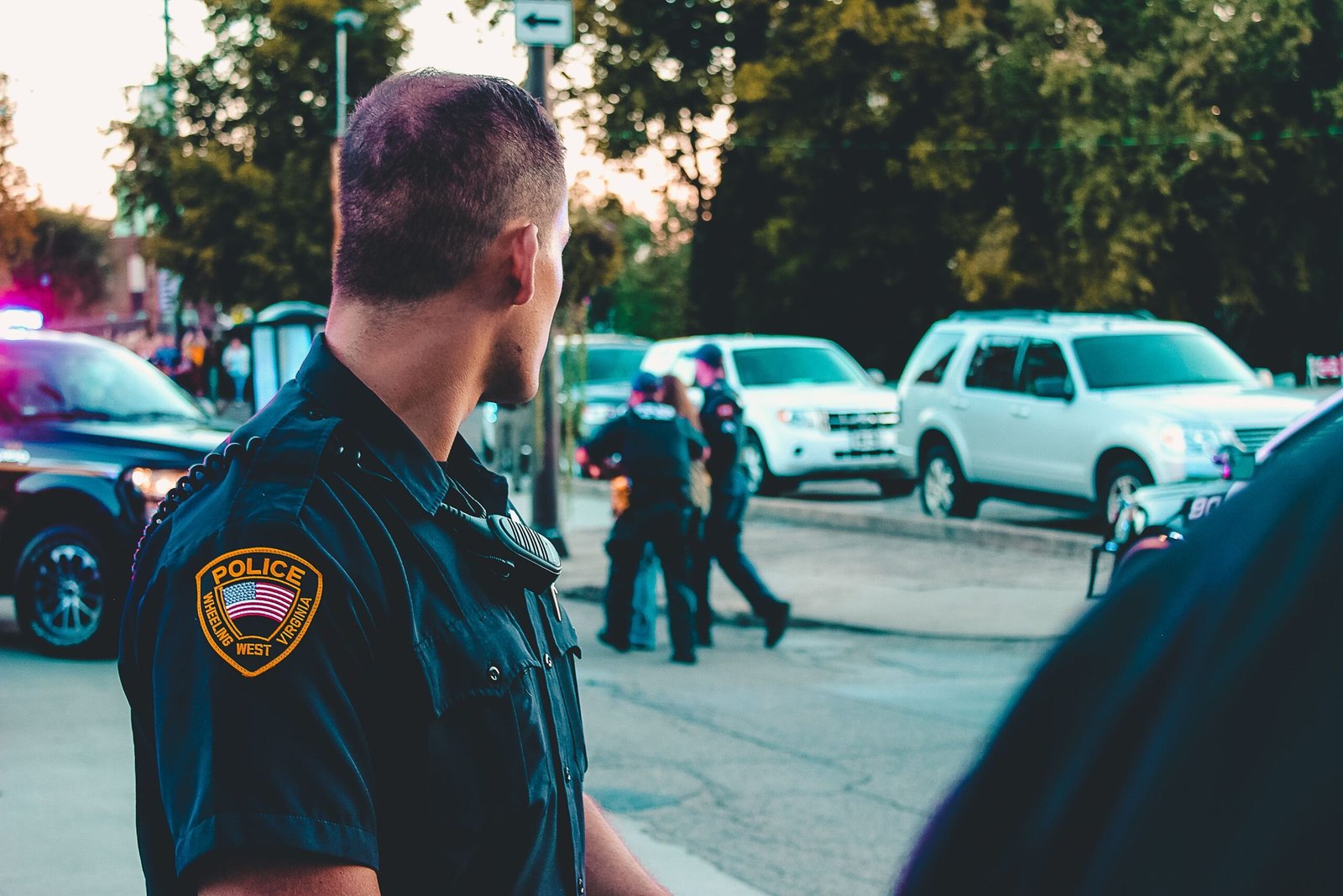 A police officer in uniform looking toward an active scene with other officers and emergency vehicles in the background.