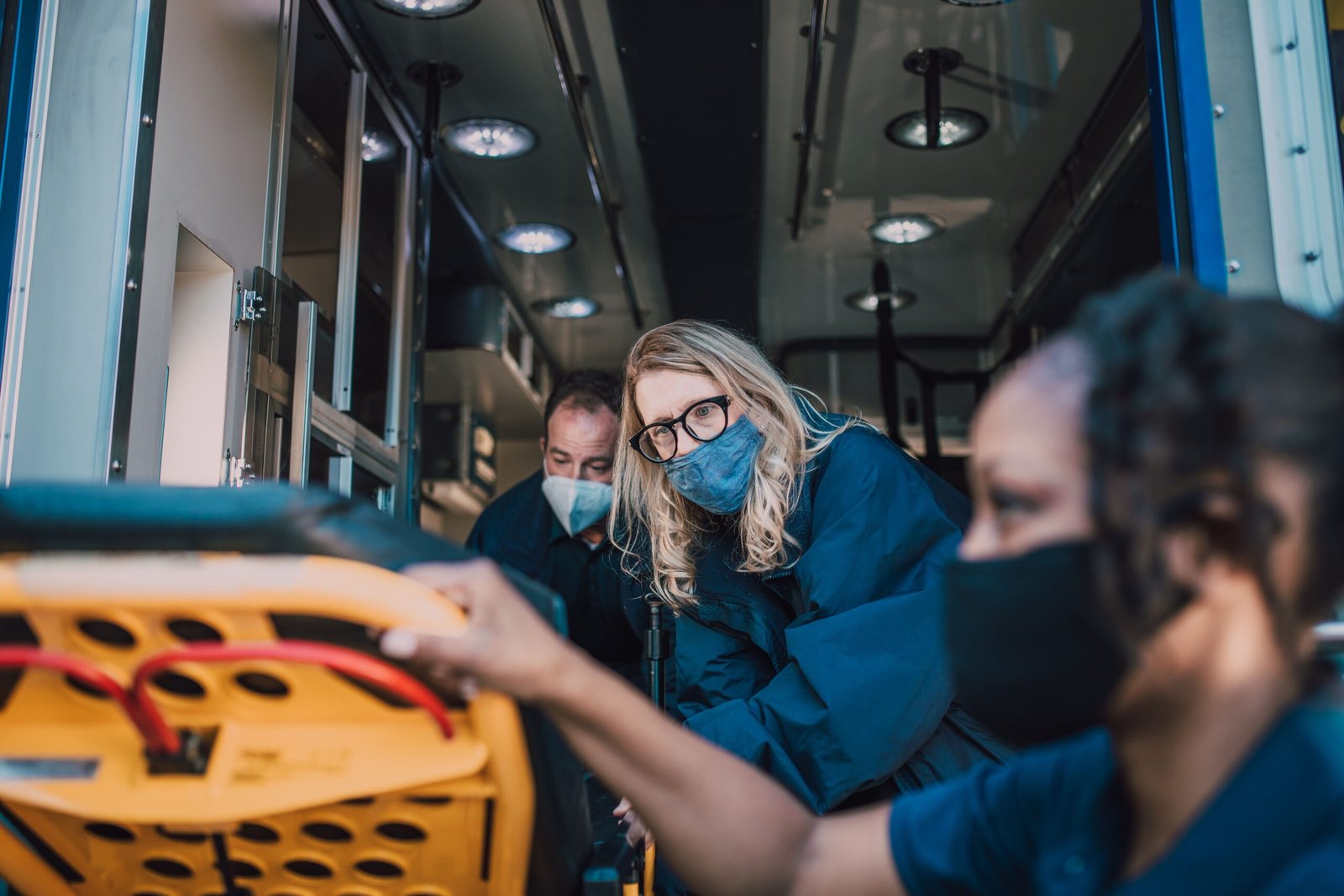 Three paramedics wearing face masks working inside the back of an ambulance, looking focused on a patient stretcher.