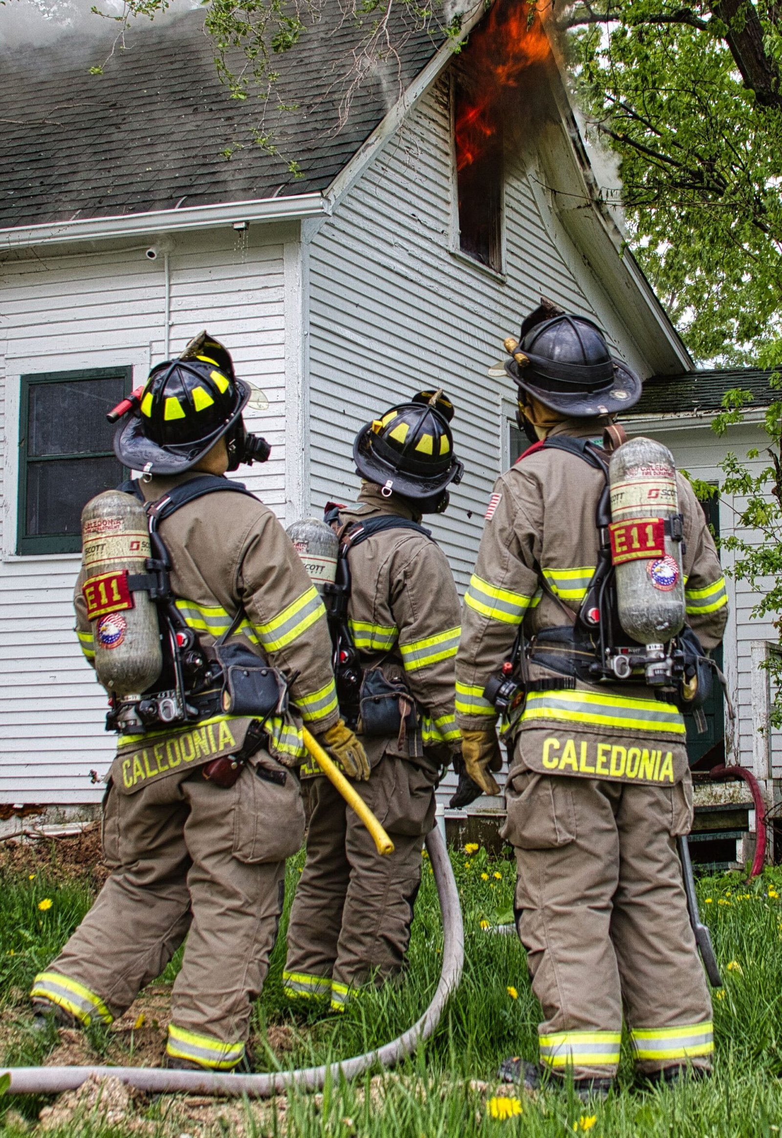Three firefighters in full protective turnout gear and helmets standing outside a white residential home with flames visible in a second-story window.