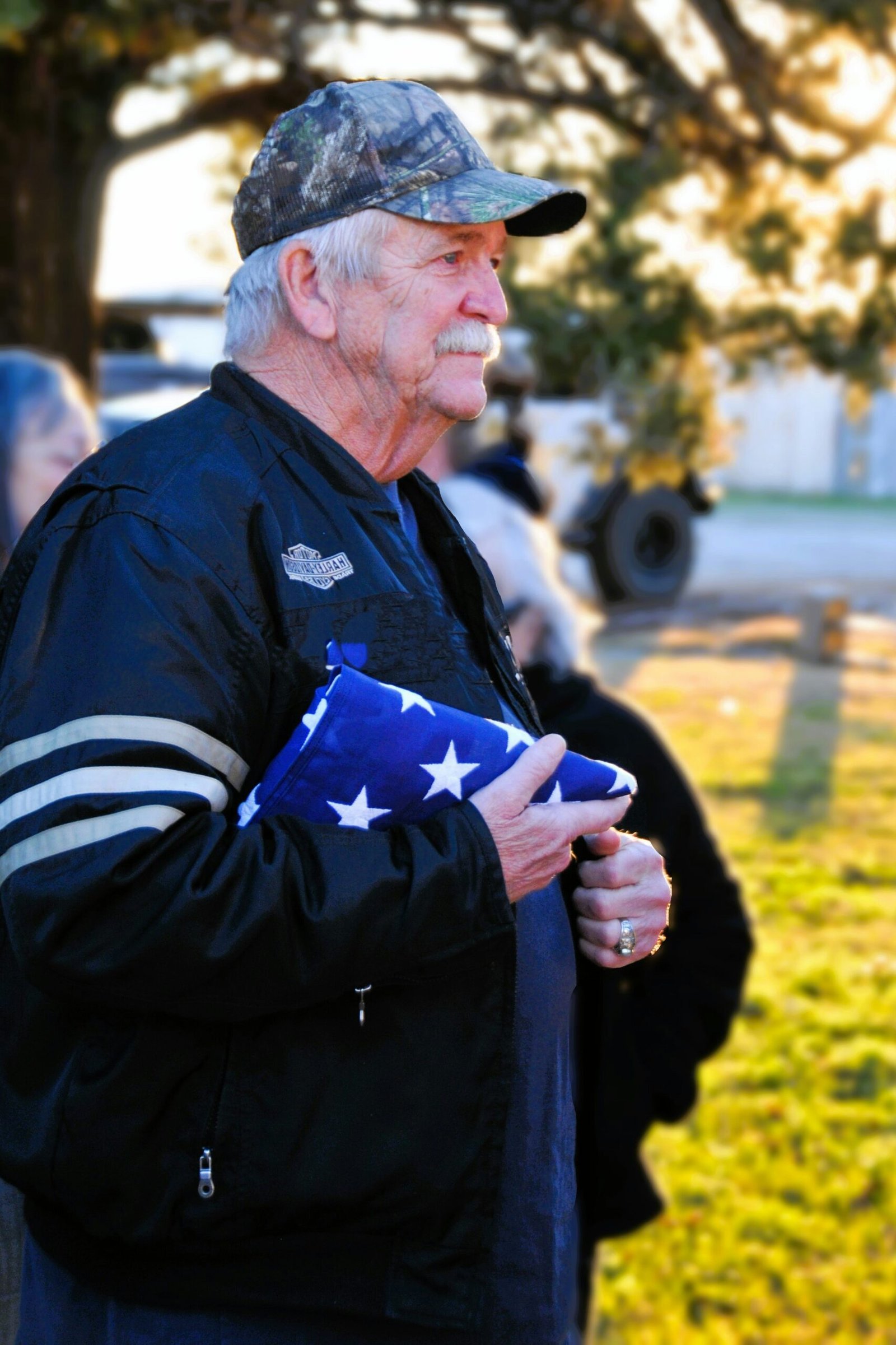 An older male veteran wearing a dark jacket and cap standing outdoors, holding a folded American flag against his chest.
