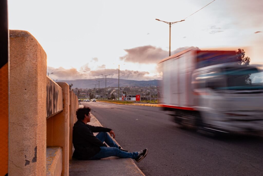 A person sitting at the roadside at dusk as traffic passes.