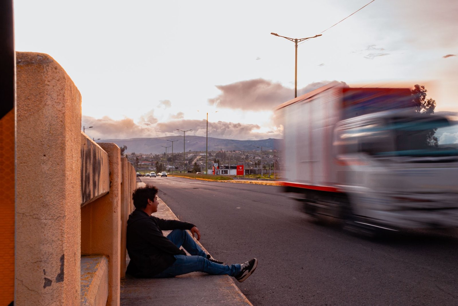 A person sitting at the roadside at dusk as traffic passes.